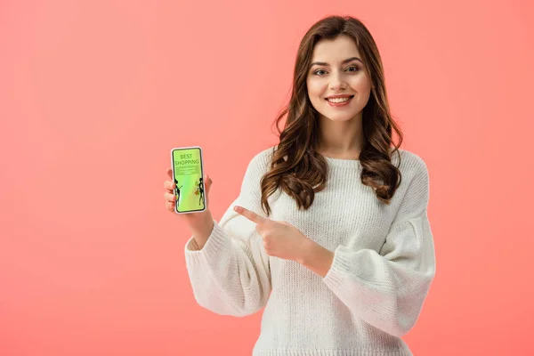 Mujer en suéter blanco apuntando con el dedo al teléfono inteligente con la mejor aplicación de compras en la pantalla aislada en rosa - foto de stock