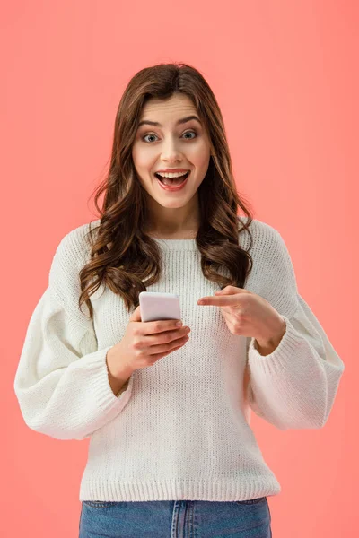 Mujer sorprendida en suéter blanco apuntando con el dedo al teléfono inteligente aislado en rosa - foto de stock