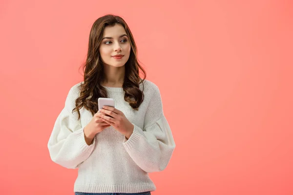 Atractiva mujer joven en suéter blanco sosteniendo teléfono inteligente aislado en rosa - foto de stock