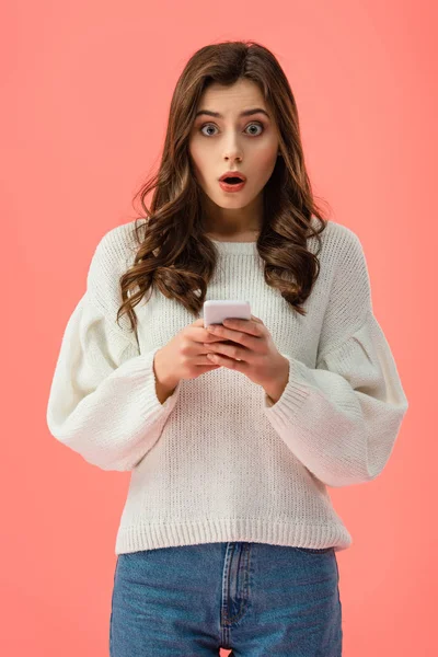 Shocked young woman in white sweater holding smartphone isolated on pink — Stock Photo