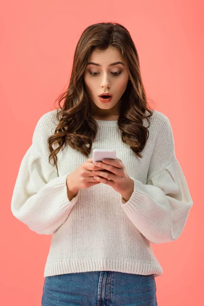 Shocked young woman in white sweater holding smartphone isolated on pink — Stock Photo