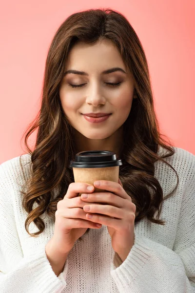 Sonriente joven con los ojos cerrados en suéter blanco sosteniendo taza de papel aislado en rosa — Stock Photo