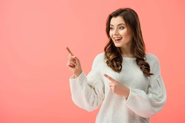 Smiling and beautiful woman in white sweater pointing with fingers and looking away isolated on pink — Stock Photo