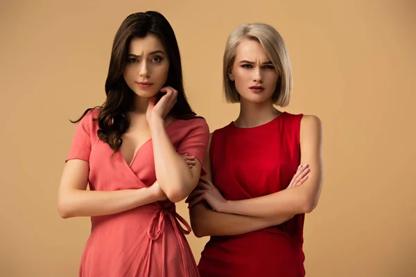 Mujeres hermosas molestas en vestidos rojos con brazos cruzados mirando a la cámara aislada en beige - foto de stock