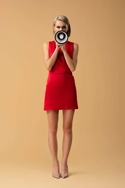 Vista completa de la mujer en vestido rojo gritando en megáfono sobre fondo beige - foto de stock