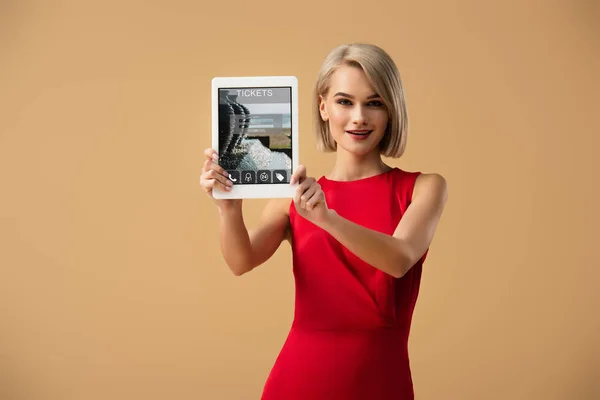Hermosa mujer en vestido rojo sosteniendo tableta digital con tickets aplicación en la pantalla aislada en beige - foto de stock