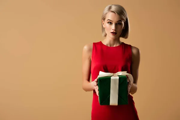 Mujer joven sorprendida y hermosa en vestido rojo sosteniendo caja de regalo aislada en beige - foto de stock