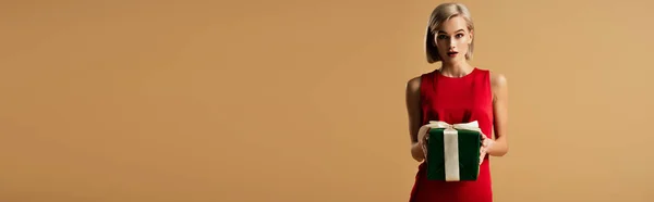 Panoramaaufnahme einer überraschten und schönen jungen Frau im roten Kleid, die eine Geschenkschachtel isoliert auf beige hält — Stockfoto