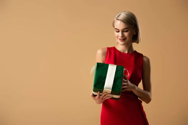 Smiling and beautiful young woman in red dress holding gift box isolated on beige — Stock Photo