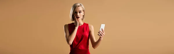 Panoramic shot of surprised young woman in red dress holding smartphone isolated on beige — Stock Photo