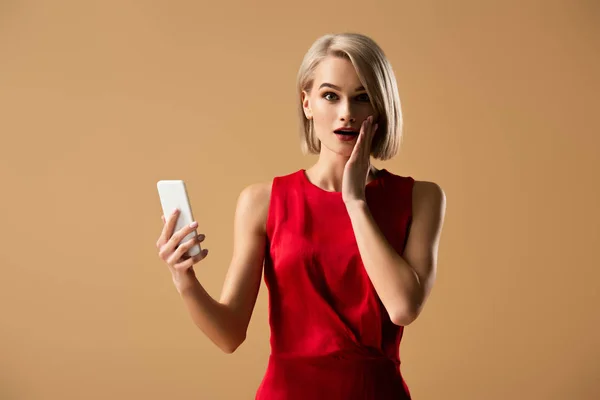Здивована молода жінка в червоній сукні тримає смартфон ізольовано на бежевому — стокове фото