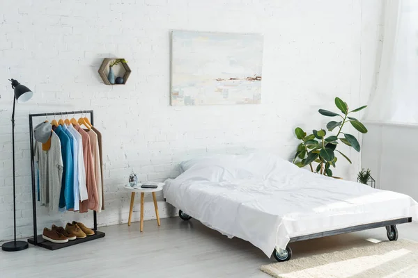 Cama, rack de roupas com camisas, planta, lâmpada de assoalho e mesa de café no quarto — Fotografia de Stock