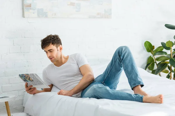 Handsome man in white t-shirt and jeans lying on bed and reading newspaper at home — Stock Photo