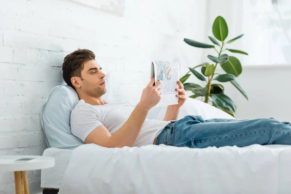 Hombre guapo en camiseta blanca y jeans acostados en la cama y leyendo el periódico en el dormitorio - foto de stock