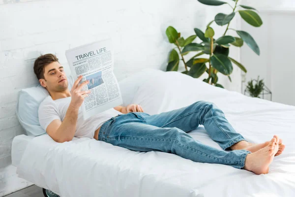 Мужчина в белой футболке и джинсах лежит на кровати и читает газету дома — стоковое фото