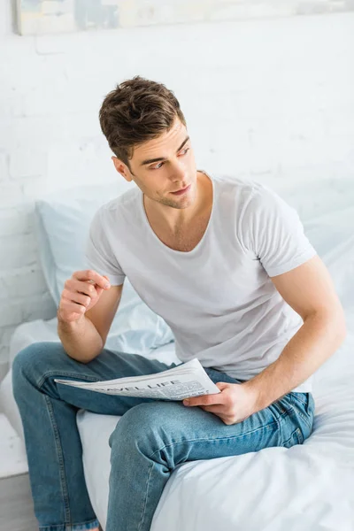 Handsome man in white t-shirt and jeans sitting on bed with newspaper at home — Stock Photo