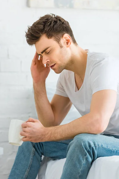 Man in white t-shirt and jeans sitting on bed, holding cup and feeling headache in bedroom — Stock Photo