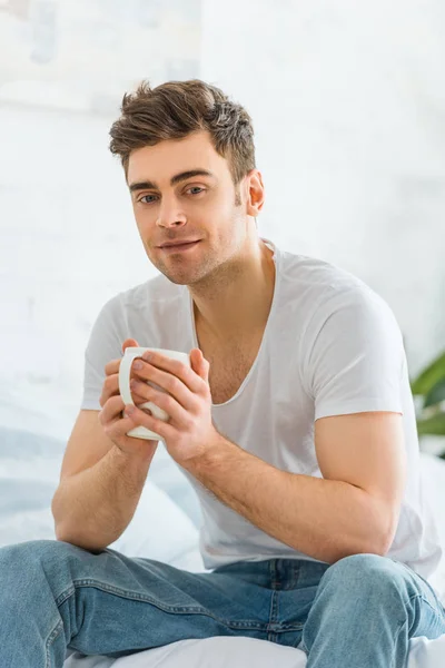 Homem bonito em t-shirt branca e jeans sentado na cama com copo no quarto — Fotografia de Stock