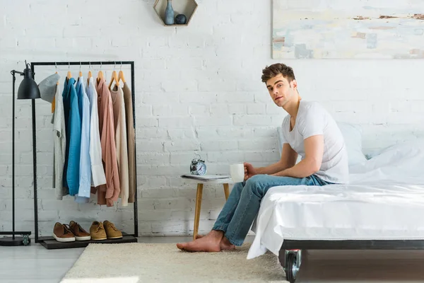 Handsome man in t-shirt and jeans sitting on bed with white cup in bedroom — Stock Photo