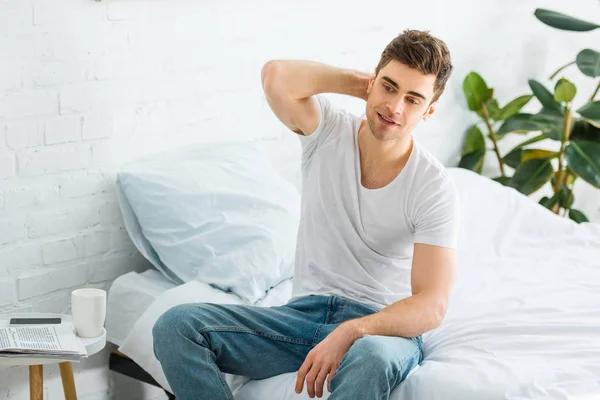 Handsome man in t-shirt and jeans sitting on bed in bedroom — Stock Photo
