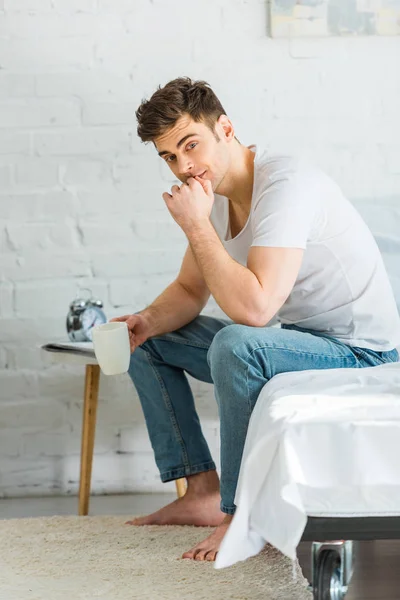 Man in white t-shirt and jeans sitting on bed and holding cup in bedroom — Stock Photo