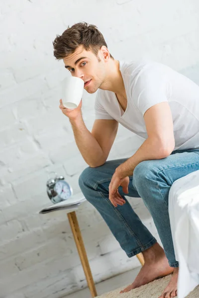 Homem de t-shirt branca e jeans sentado na cama e beber café no quarto — Fotografia de Stock