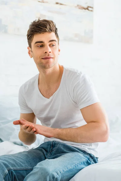 Handsome man in white t-shirt and jeans sitting on bed at home — Stock Photo