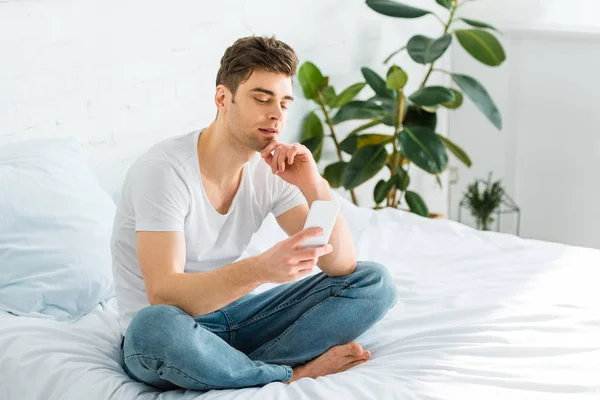Красивый мужчина в белой футболке и джинсах сидит на кровати со смартфоном дома — стоковое фото