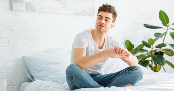 Panoramic shot of handsome man in white t-shirt and jeans sitting on bed at home — Stock Photo