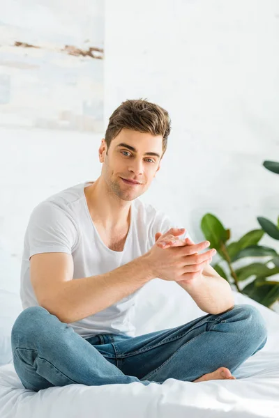 Selective focus of handsome man in white t-shirt and jeans sitting on bed at home — Stock Photo