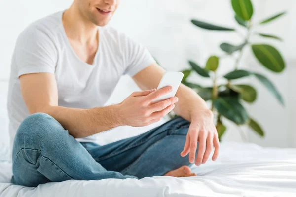 Cropped view of man in white t-shirt and jeans sitting on bed with smartphone at home — Stock Photo