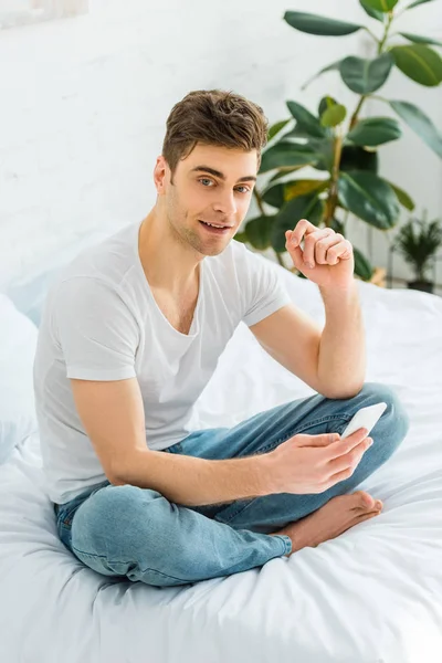 Handsome man in white t-shirt and jeans sitting on bed and holding smartphone at home — Stock Photo