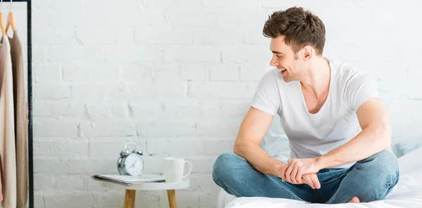 Colpo panoramico di bell'uomo in t-shirt bianca e jeans seduto sul letto e sorridente a casa — Foto stock