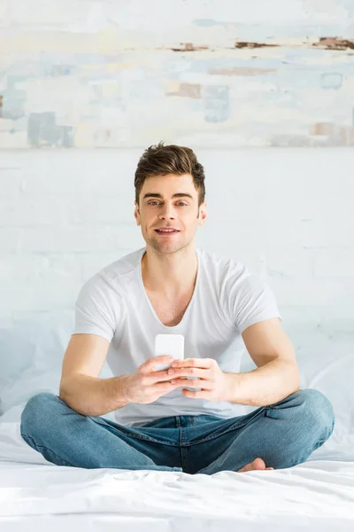 Hombre guapo en camiseta y jeans sentado en la cama y sosteniendo el teléfono inteligente en el dormitorio - foto de stock