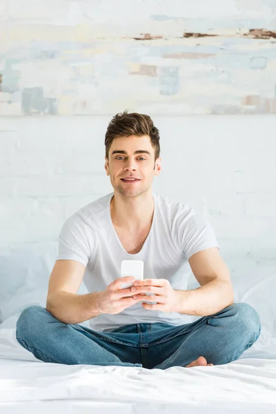 Bel homme en t-shirt et jeans assis sur le lit et tenant smartphone dans la chambre — Photo de stock