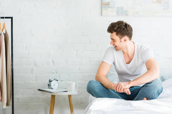 Bel homme en t-shirt blanc et jeans assis sur le lit et souriant à la maison — Photo de stock