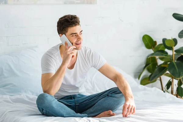 Hombre en camiseta y jeans sentado en la cama y hablando en el teléfono inteligente en el dormitorio - foto de stock