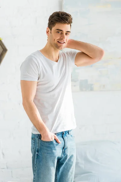 Homem bonito em t-shirt branca e jeans de pé perto da cama no quarto — Fotografia de Stock