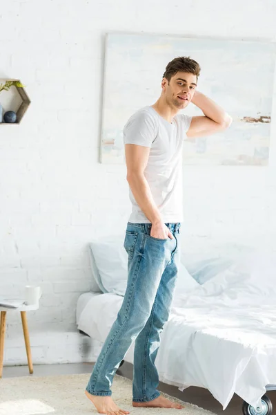 Bel homme en t-shirt blanc et jeans debout près du lit à la maison — Photo de stock