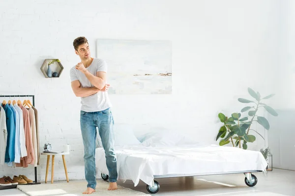 Uomo in t-shirt e jeans in piedi vicino a letto e appendiabiti in camera da letto — Foto stock