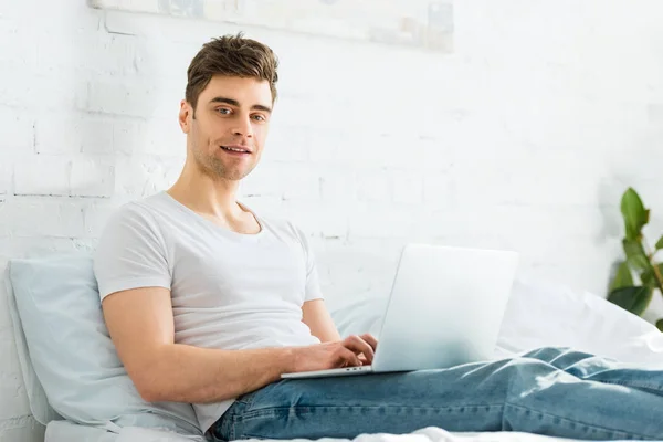 Hombre guapo en camiseta blanca y jeans sentados en la cama y escribiendo en el ordenador portátil en el dormitorio - foto de stock