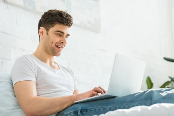 Hombre guapo en camiseta blanca y jeans sentados en la cama, sonriendo y escribiendo en el ordenador portátil en el dormitorio - foto de stock