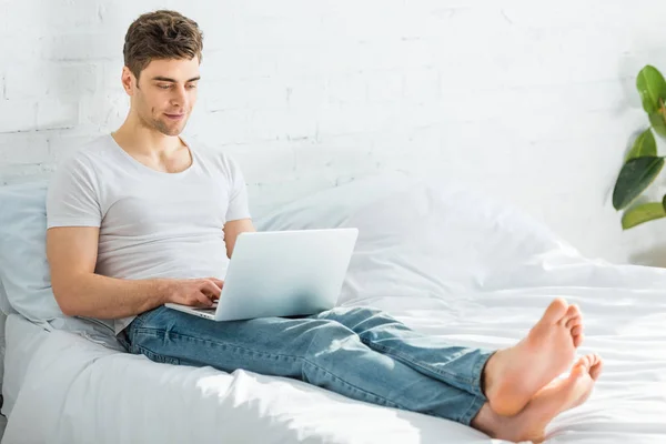 Hombre guapo en camiseta blanca y jeans sentados en la cama y escribiendo en el ordenador portátil en el dormitorio - foto de stock