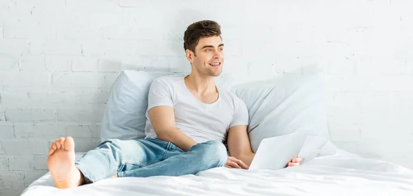 Панорамный снимок красивого мужчины в белой футболке и джинсах, сидящего на кровати с ноутбуком в спальне — стоковое фото
