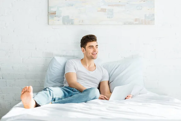 Hombre guapo en camiseta blanca y vaqueros sentados en la cama con el ordenador portátil y sonriendo en el dormitorio - foto de stock