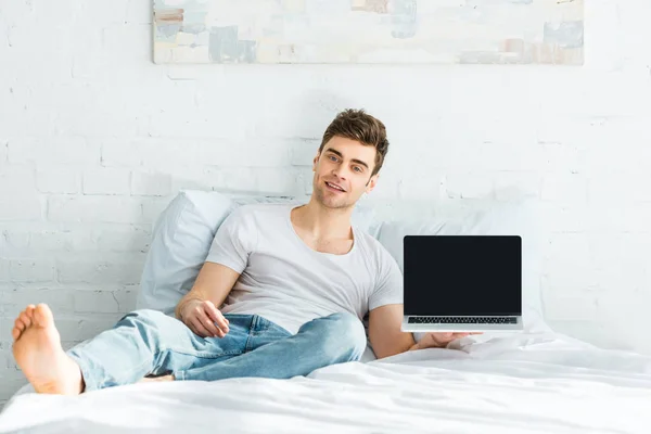 Handsome man in white t-shirt and jeans sitting on bed and holding laptop with blank screen in bedroom — Stock Photo