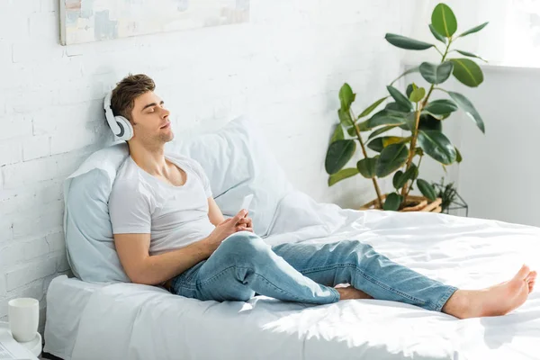 Bel homme en t-shirt blanc et jeans assis sur le lit, tenant smartphone et écoutant de la musique dans la chambre — Photo de stock