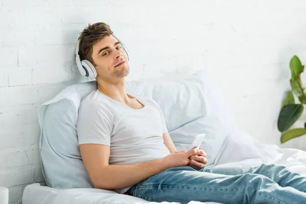 Man in white t-shirt and jeans sitting on bed, holding smartphone and listening music in bedroom — Stock Photo