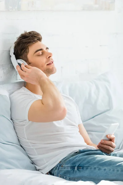 Homem bonito em camiseta branca com olhos fechados sentado na cama, segurando smartphone e ouvindo música no quarto — Fotografia de Stock