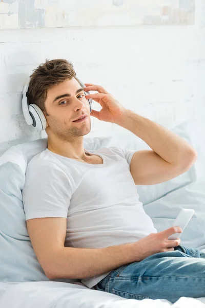 Man in white t-shirt and jeans sitting on bed with smartphone and listening music in bedroom — Stock Photo